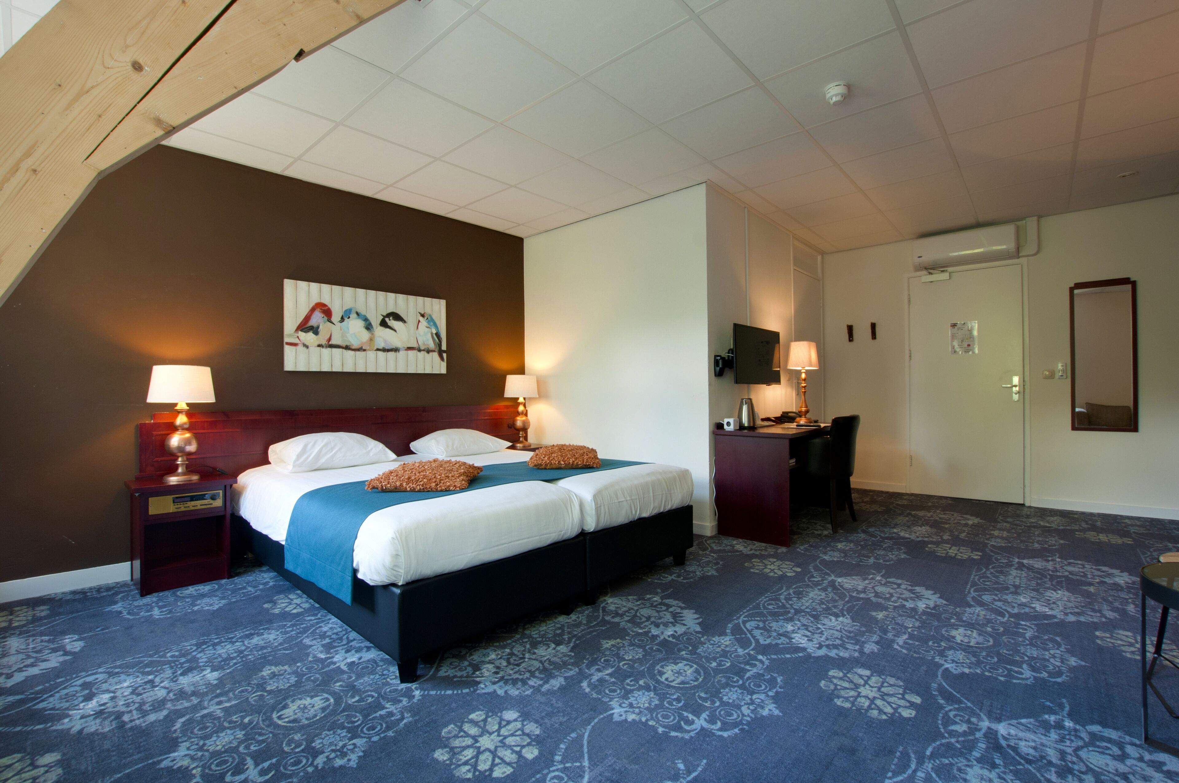 Een hotelkamer met een groot bed, twee bedlampen, een bureau met stoel, een televisie en een schilderij aan de muur.