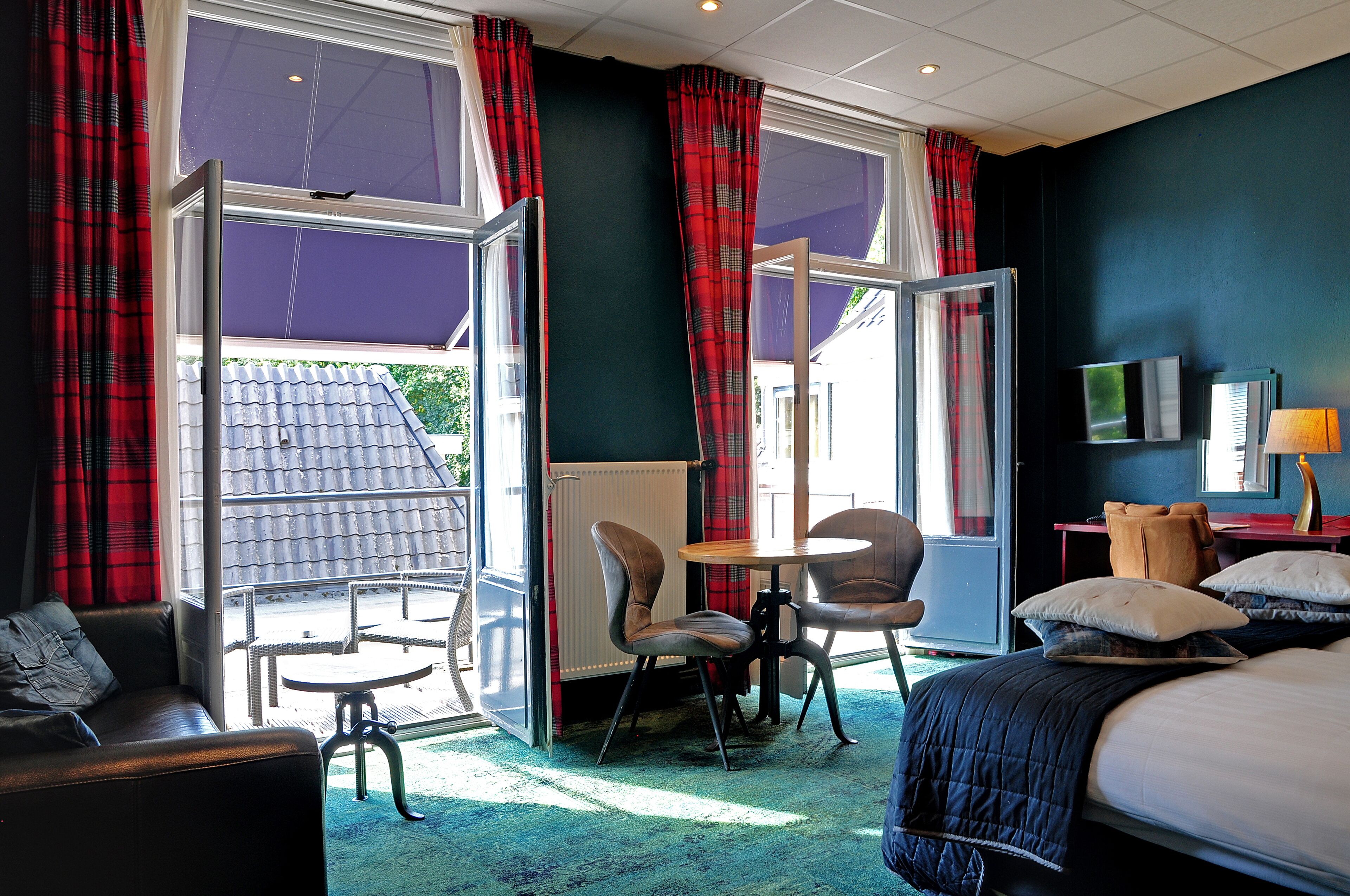 A hotel room with a bed, a sofa, a table, and chairs. There are curtains on the windows, and a radiator is visible.