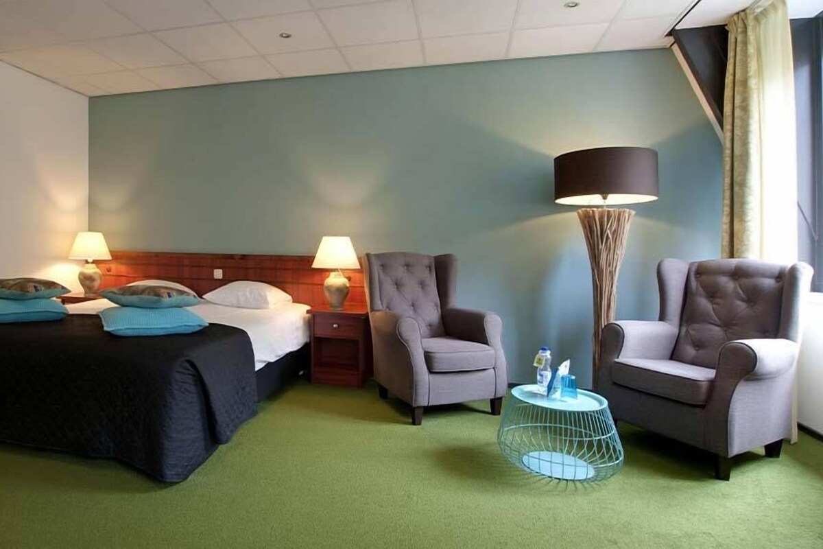 A hotel room with a bed, two armchairs, a side table, and a floor lamp.