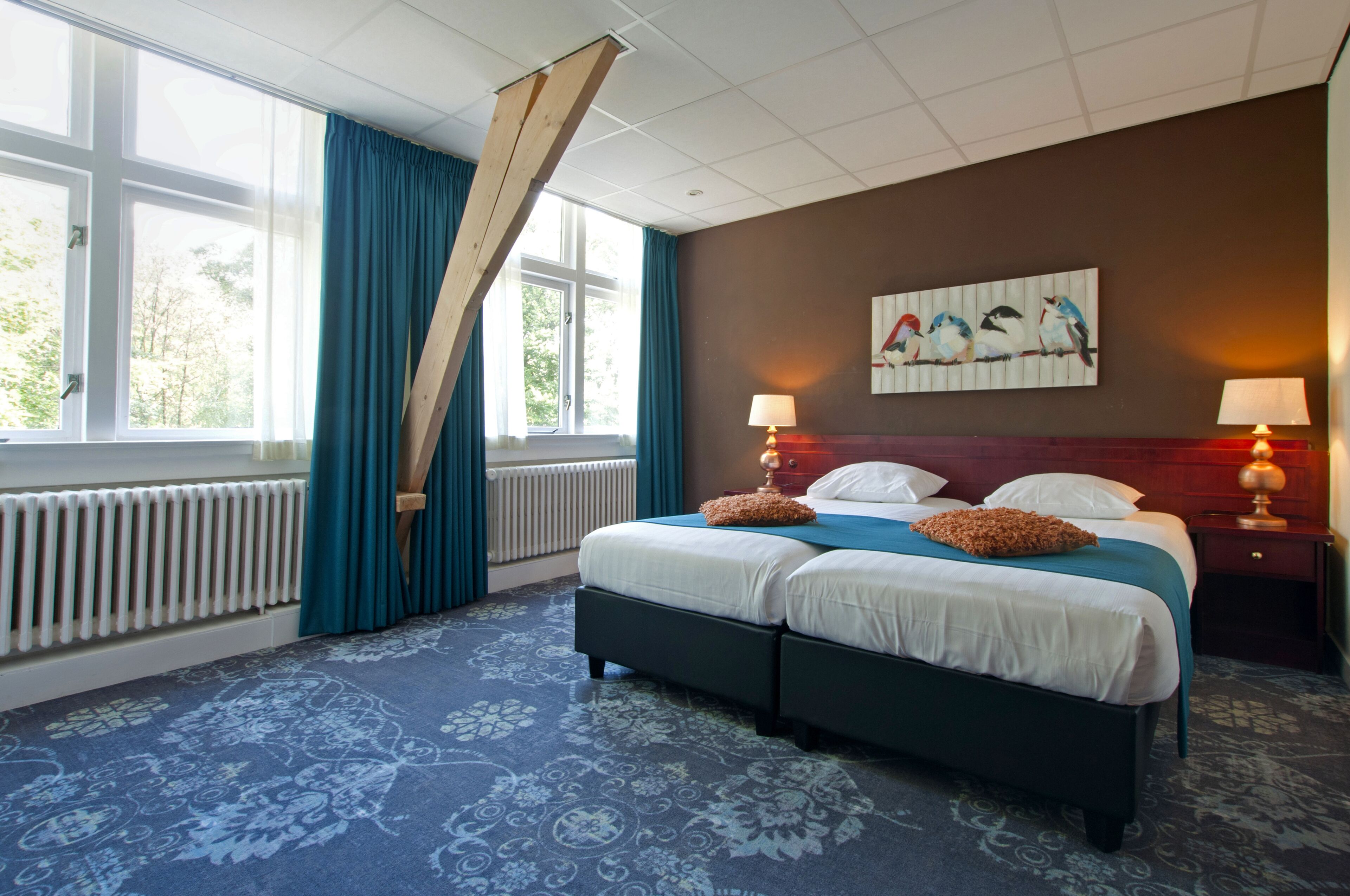 A double bed with two pillows, a blue and white patterned carpet, a wooden headboard, a painting of birds on the wall, and two bedside lamps.