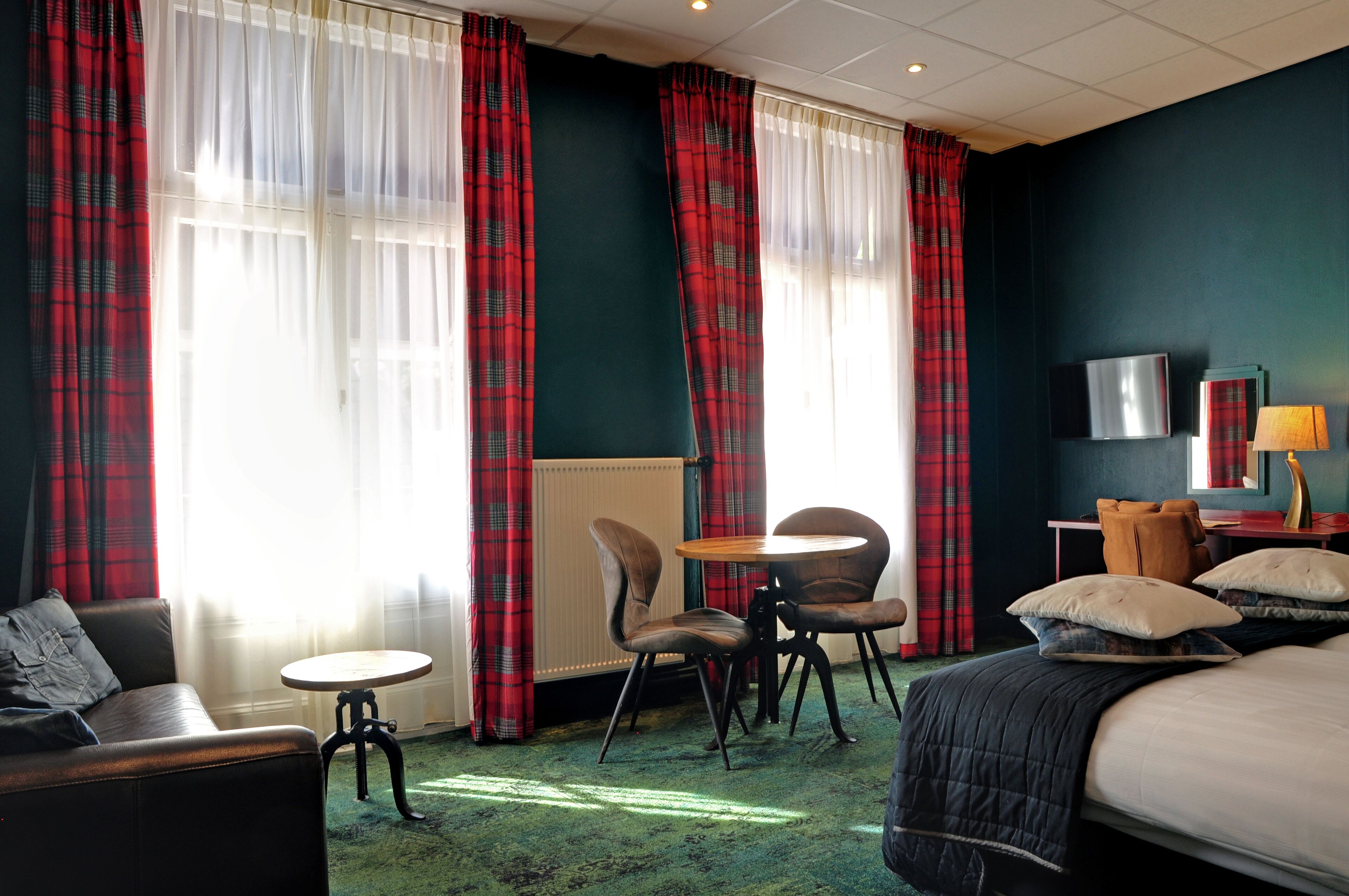 Habitación de hotel con cama, dos sillas, una mesita, televisor y cortinas rojas a cuadros.
