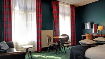 Habitación de hotel con cama, dos sillas, una mesita, televisor y cortinas rojas a cuadros.