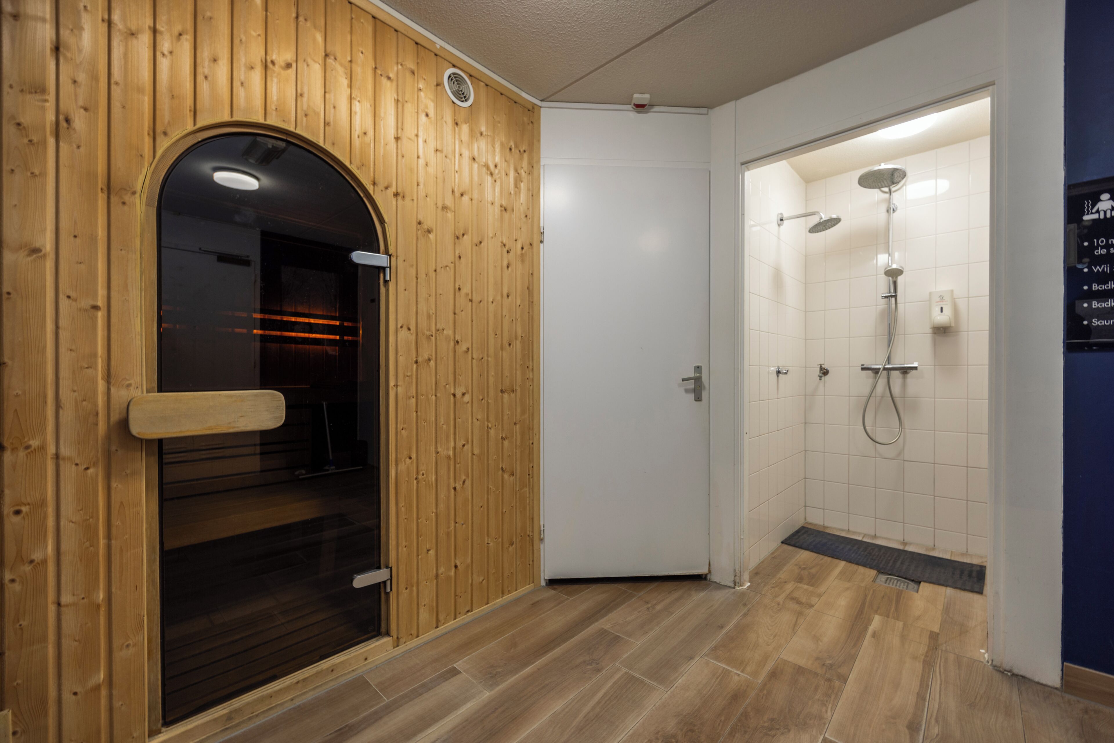 A wooden door leading to a sauna room, a white tiled shower area, and a wooden floor.