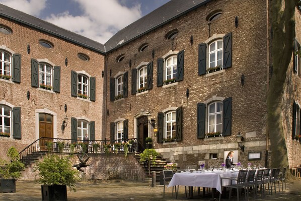 Un patio con zona de comedor preparada para un evento, con un edificio de ladrillo y múltiples ventanas, y una escalera que conduce al nivel superior.