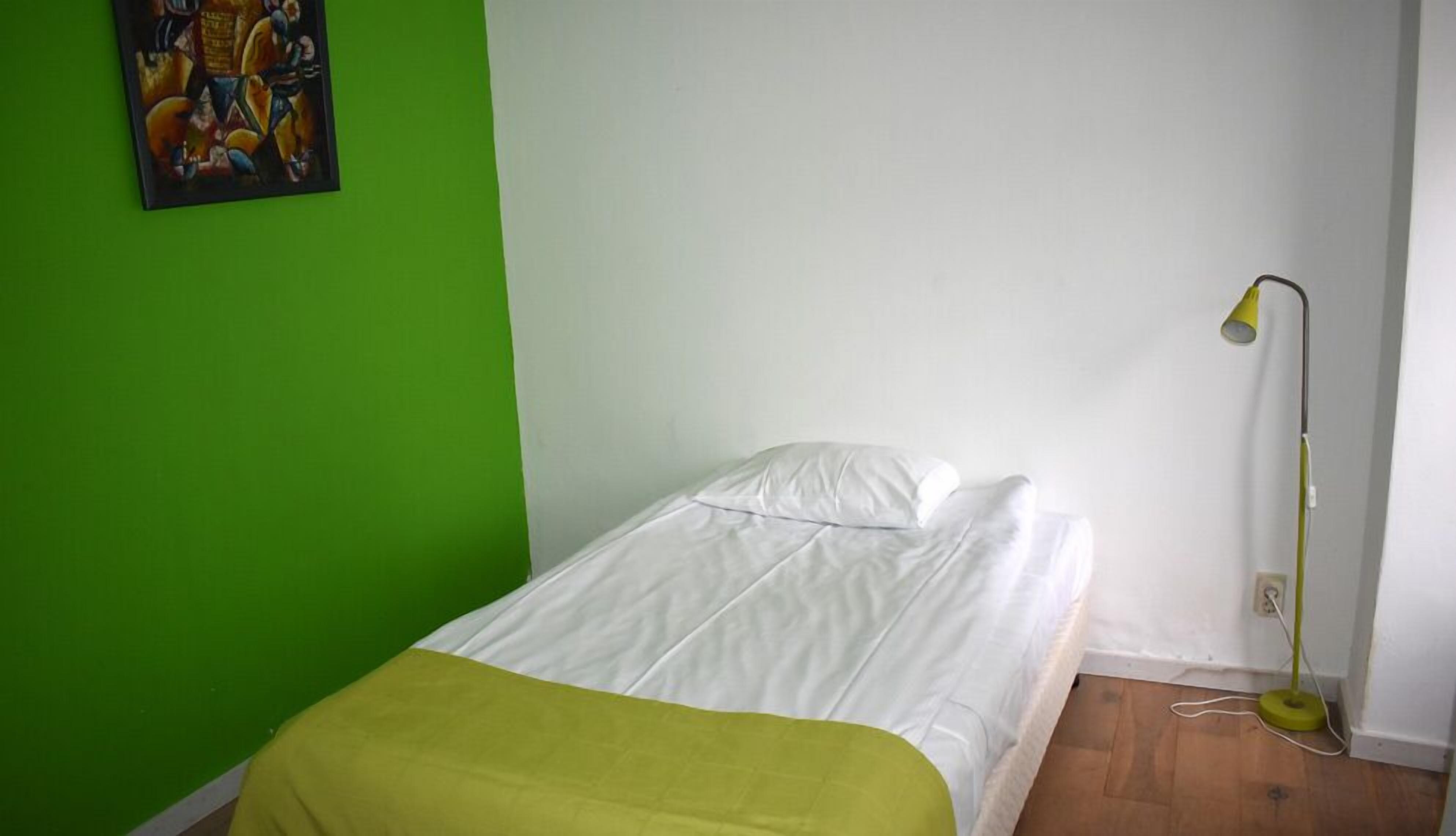 Een kleine eenpersoonskamer met een groene muur, een wit bed en een vloerlamp.