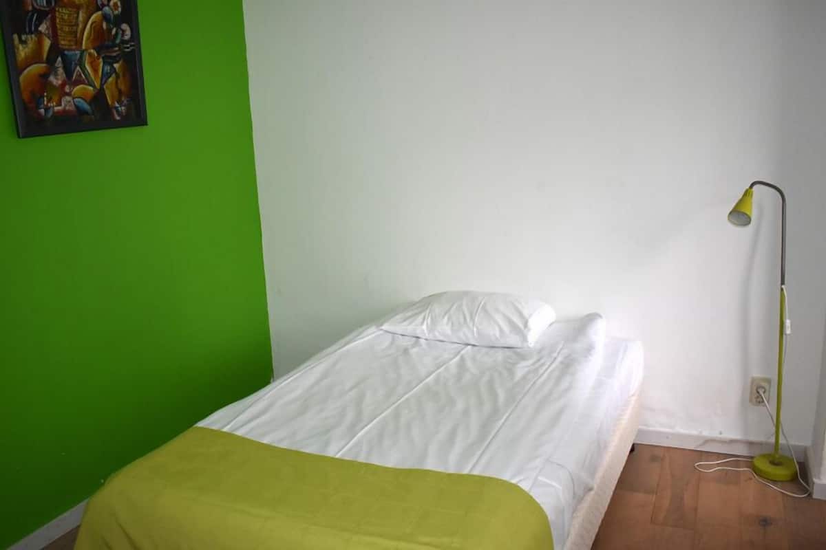 Ein kleines Zimmer mit einem Einzelbett, einer grünen Wand, einem weißen Bett und einer Stehlampe.