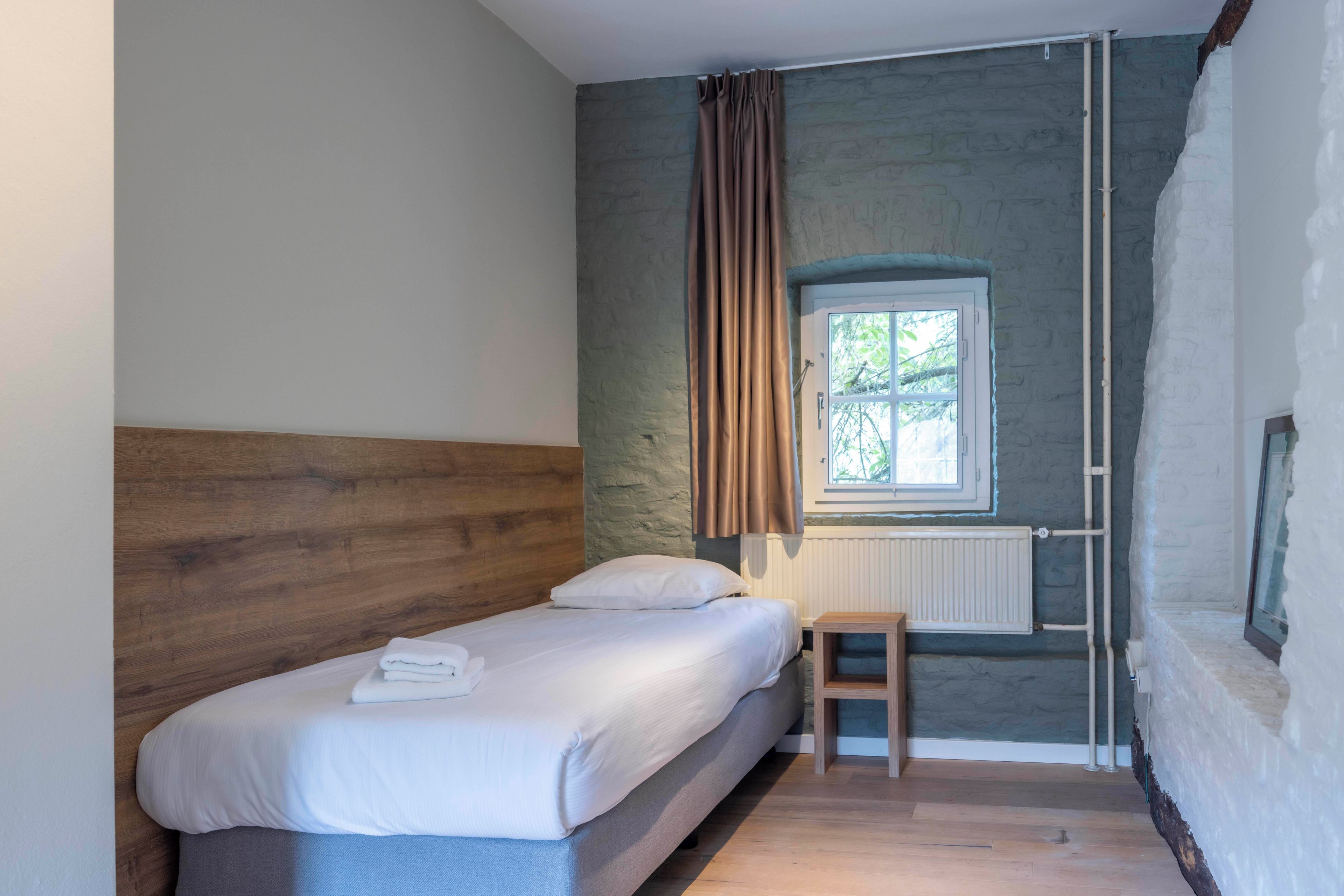 A bedroom with a wooden headboard, a single bed with white linens, a small wooden stool, a window with curtains, and a radiator.
