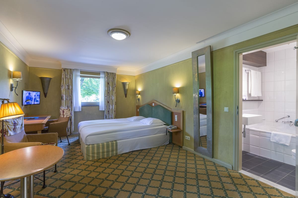 Une chambre d’hôtel avec un lit double, un bureau avec une chaise, une télévision, une lampe et une salle de bain avec une baignoire et un lavabo.