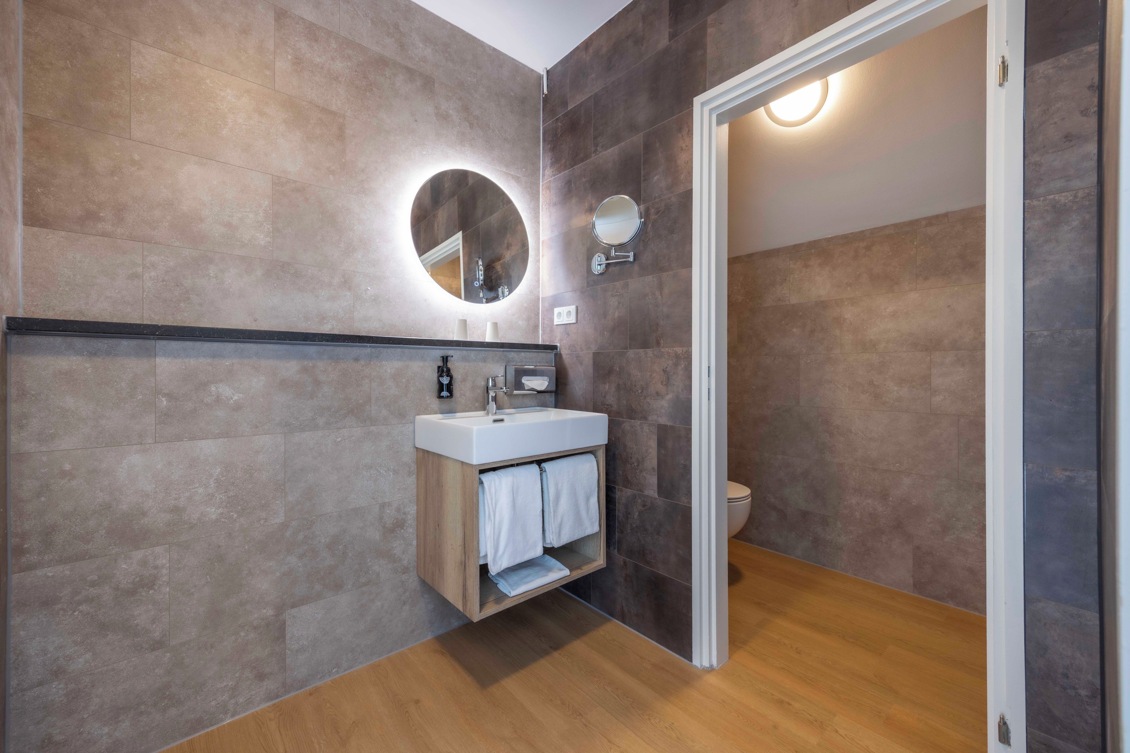 A modern bathroom with a wooden vanity, a round mirror, and a toilet.