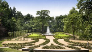 Een aangelegde tuin met een centrale fontein, omringd door keurig bijgesnoeid groen en bomen.