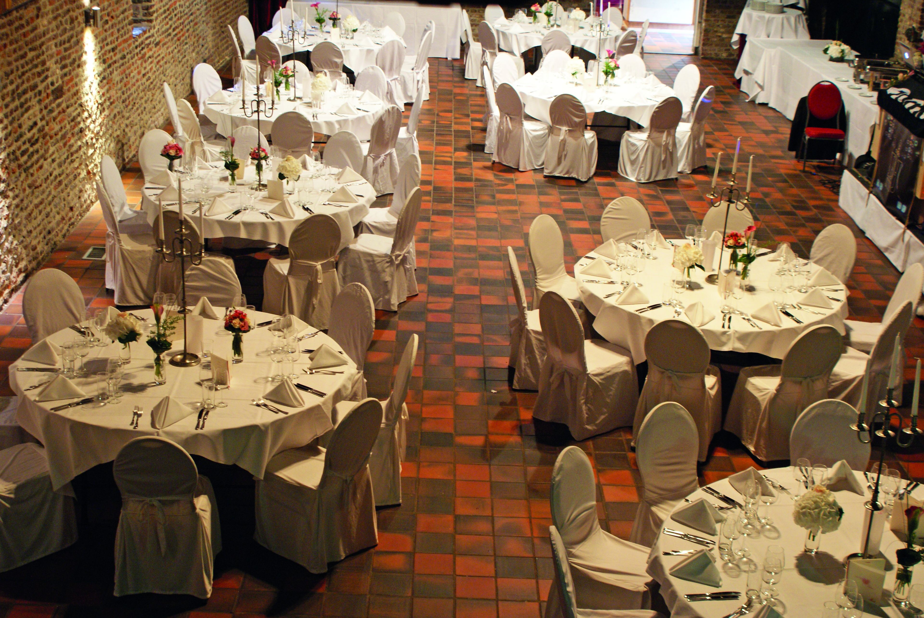 Une salle de banquet avec des tables rondes dressées pour le repas, chacune étant recouverte d’une nappe blanche et agrémentée de chaises et de compositions florales.