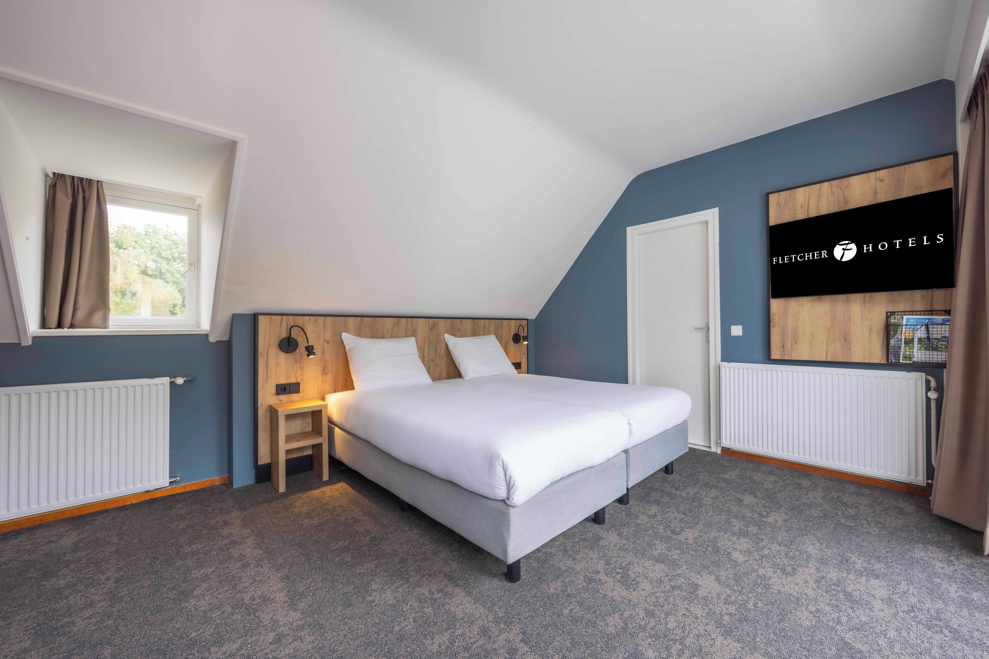 A hotel room with a bed, bedside table, lamp, window with curtains, and a wall-mounted sign.