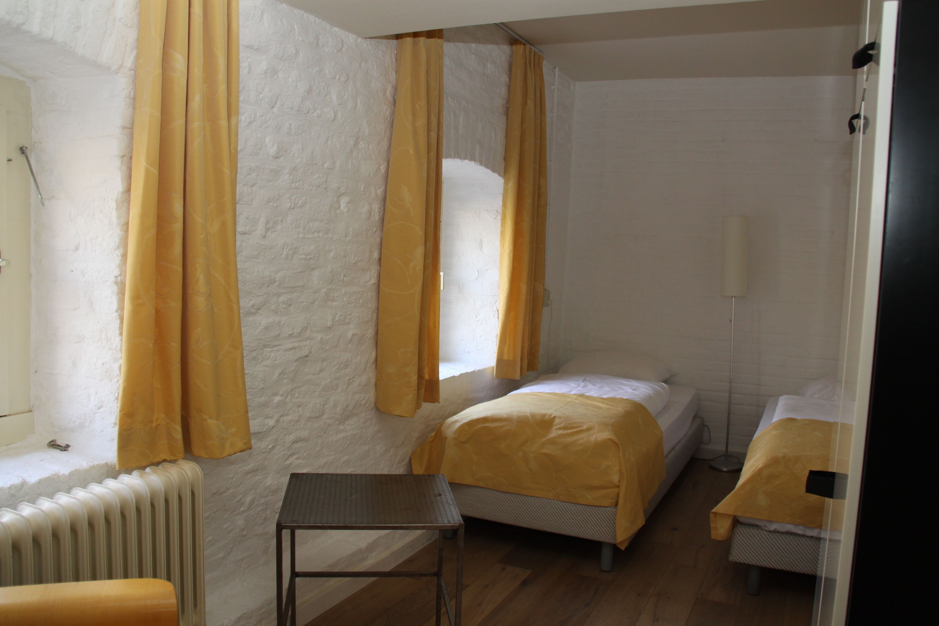 A small, bright room with two beds, a radiator, a table, and curtains.