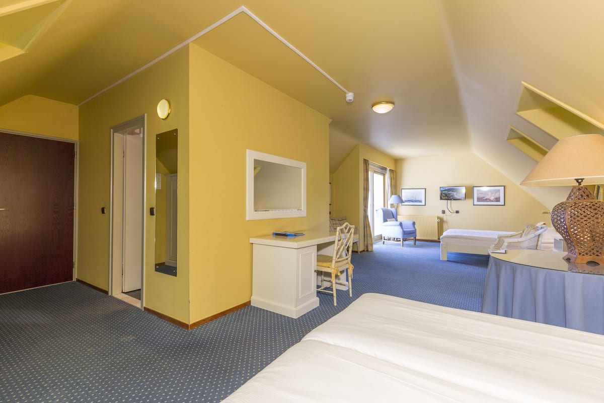 Une chambre d’hôtel avec un lit, un bureau, une chaise et une petite table avec une lampe. Il y a une porte qui donne sur une autre pièce et une fenêtre avec des rideaux.