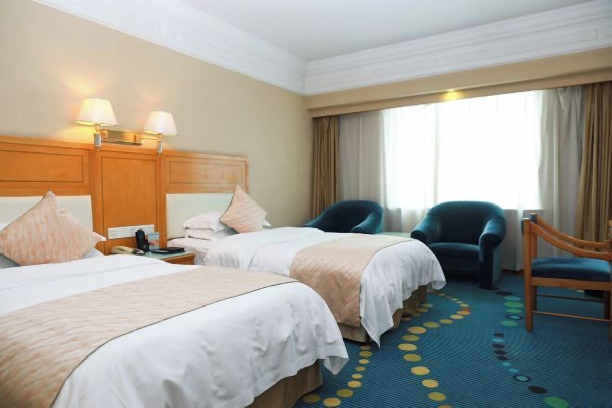 Habitación de hotel con dos camas, un escritorio, una silla y un amplio ventanal con cortinas.