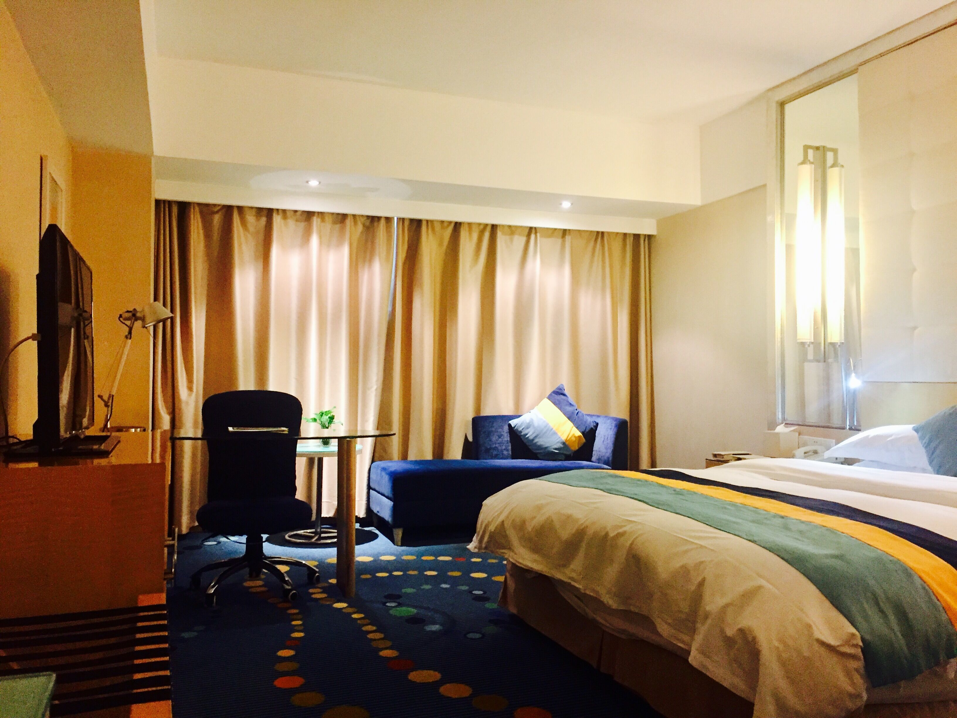 A hotel room with a bed, a desk, a chair, a small table with a plant, and curtains.