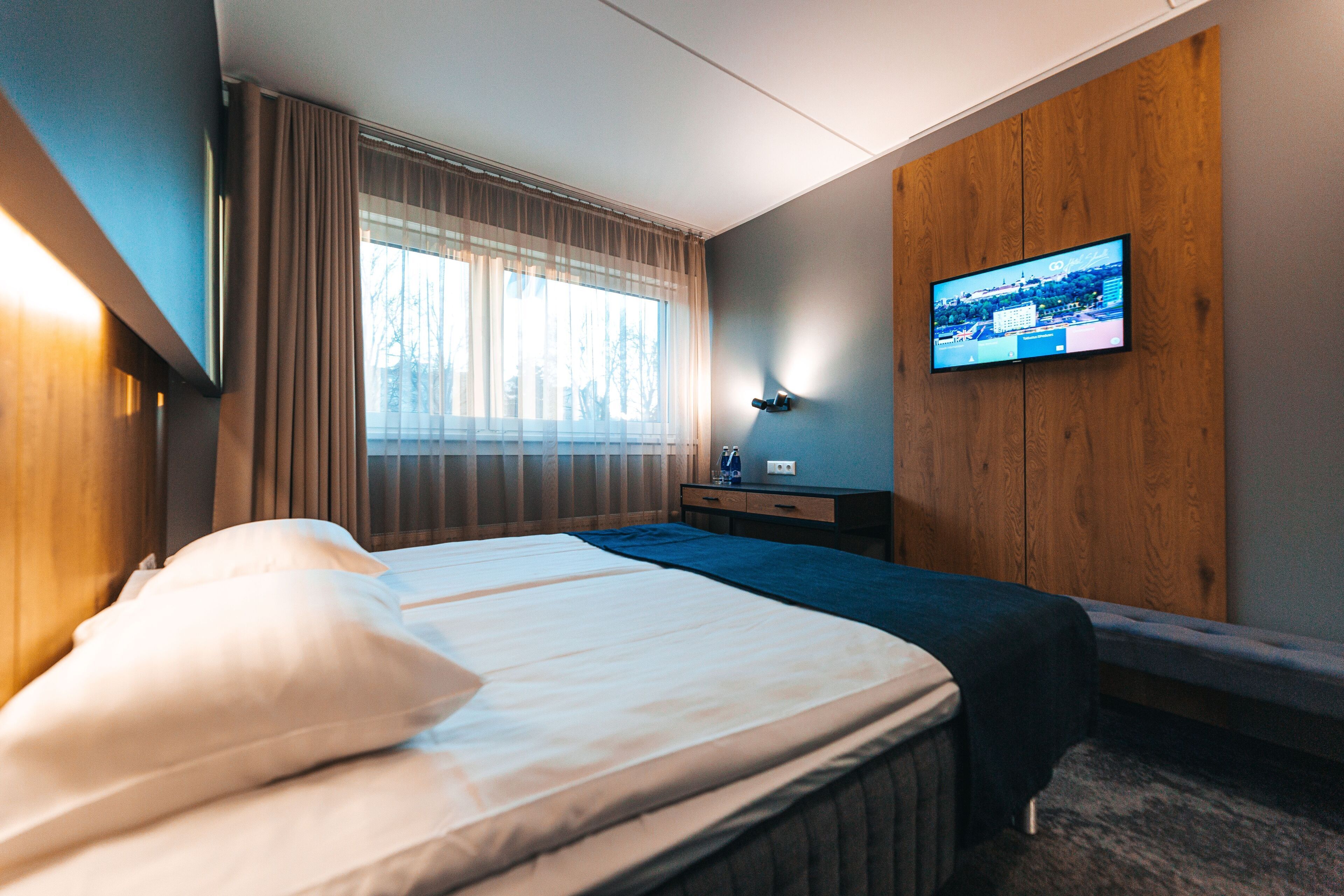 Habitación de hotel con cama, televisión montada en la pared, ventana con cortinas y un panel de madera en la pared.