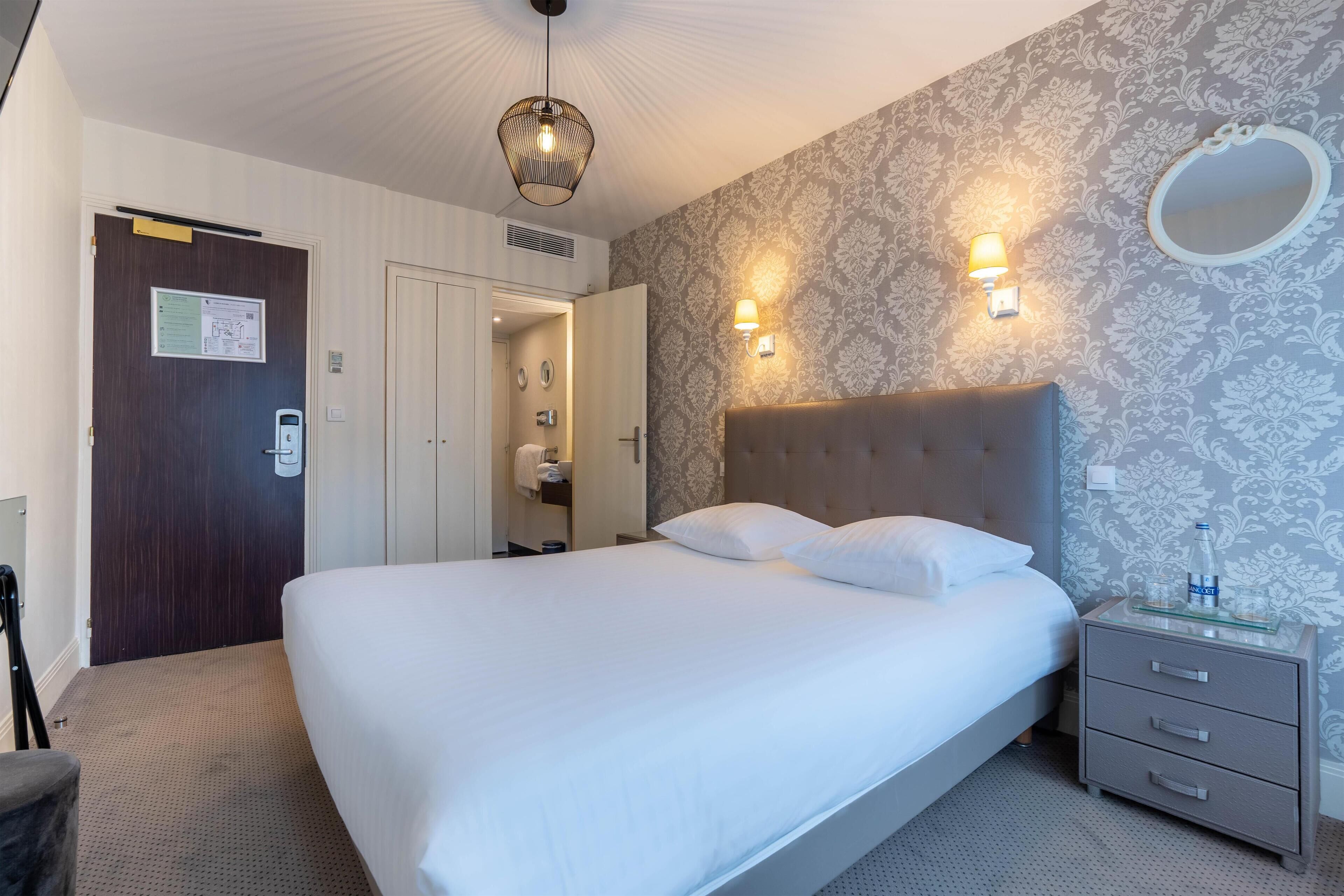 Habitación de hotel con una cama grande, una mesita de noche con una botella, un espejo y una pared con papel tapiz estampado.