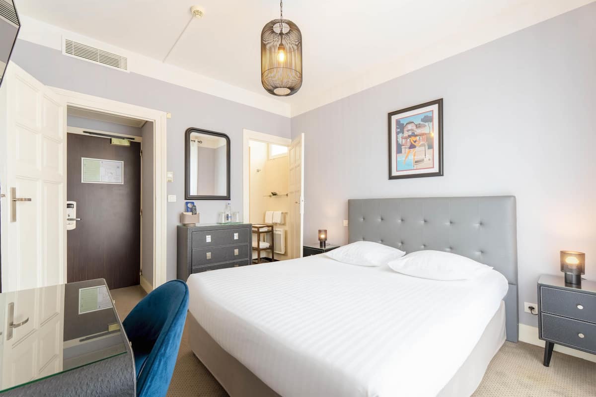 A hotel room with a large bed, a desk with a computer, a chair, a dresser, a bedside table with a lamp, and a framed picture on the wall.