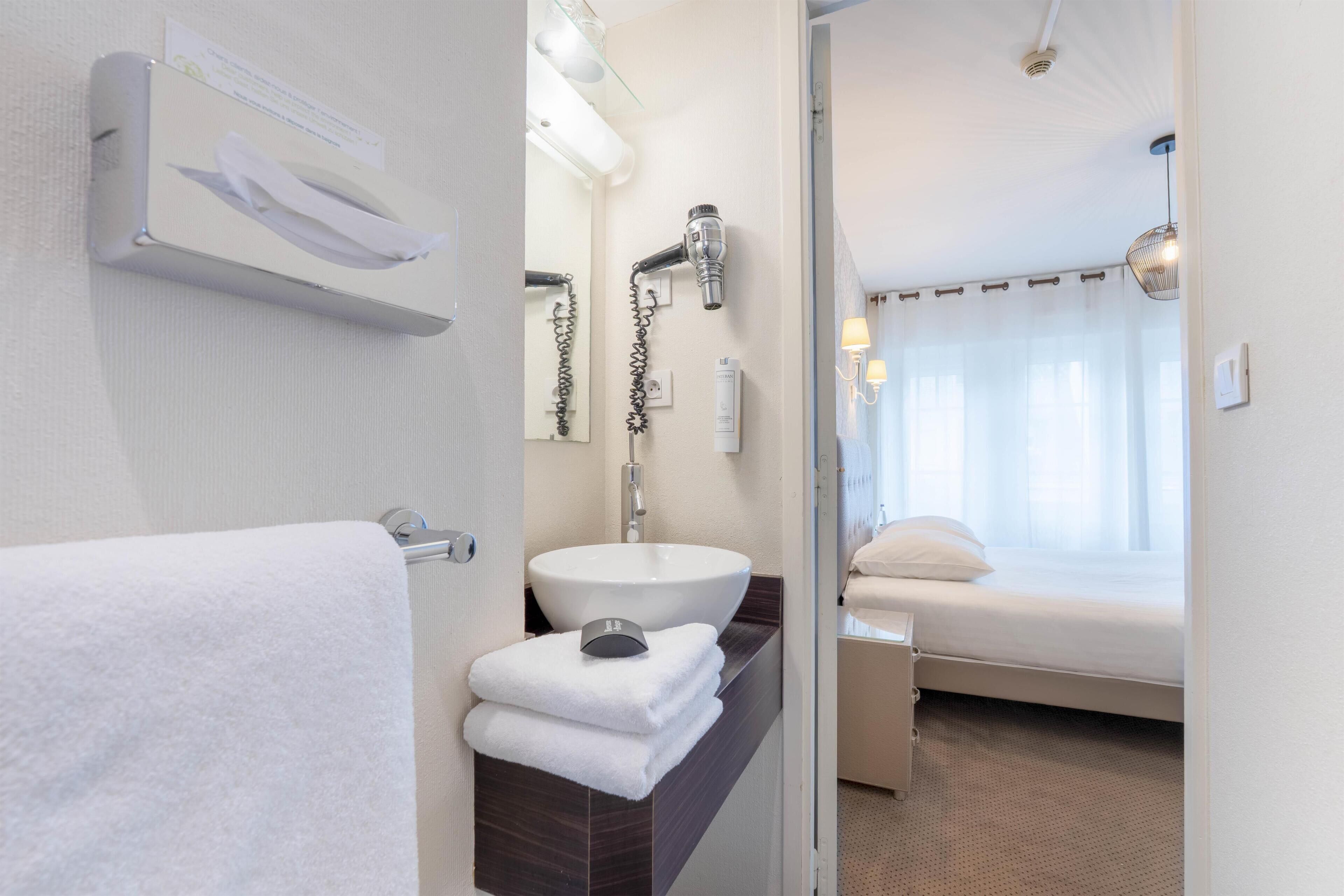 Een hotelbadkamer met een wastafel, zeepdispenser en een bed dat door een open deur zichtbaar is.