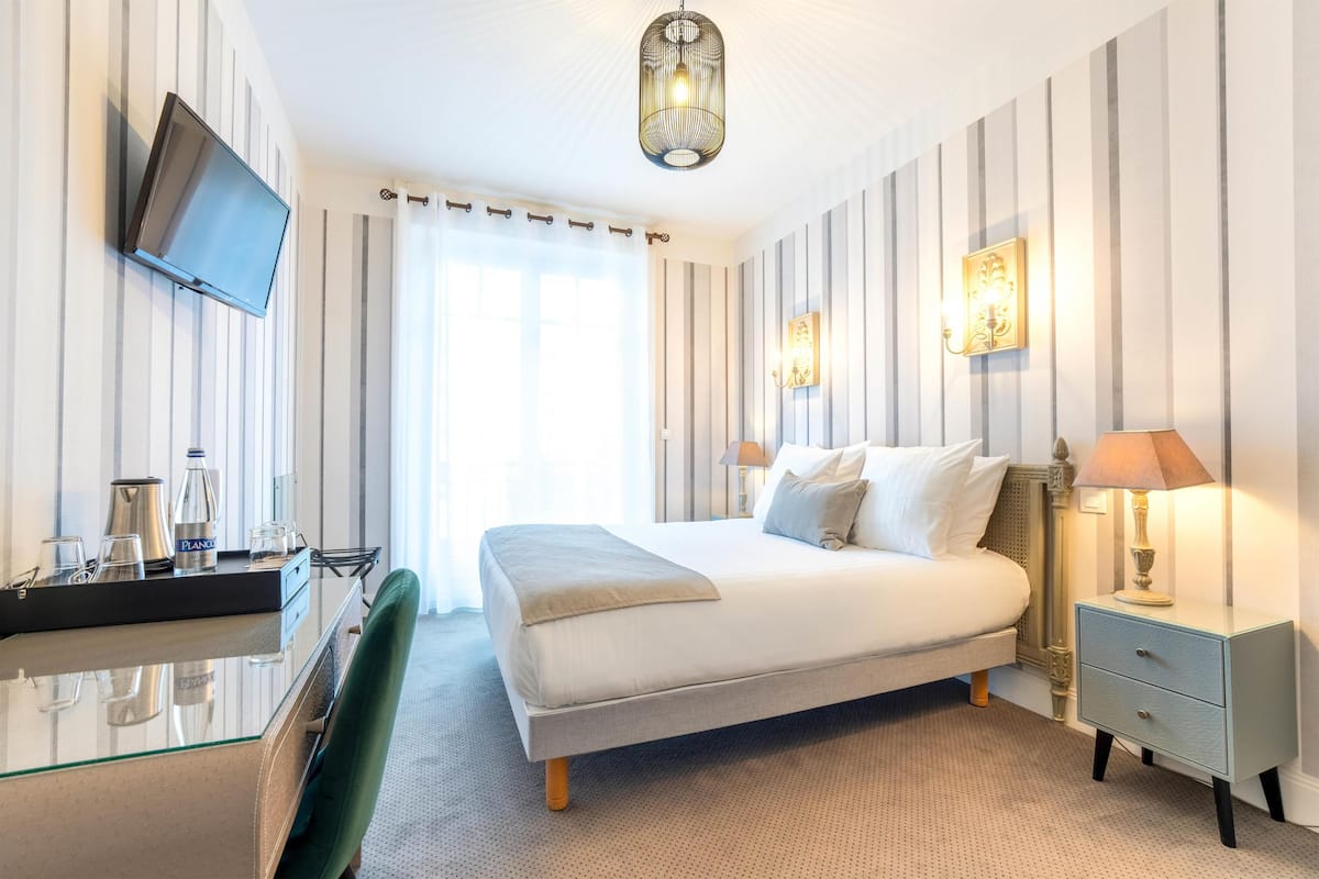 A hotel room with a bed, bedside table, lamp, TV, and a desk with a chair.