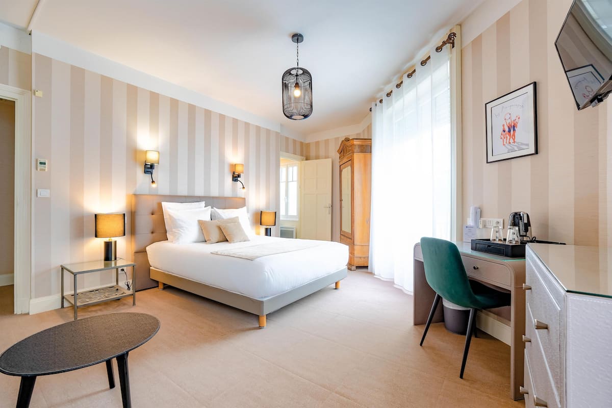 A hotel room with a bed, bedside lamps, a desk with a chair, a small table, and a wardrobe.