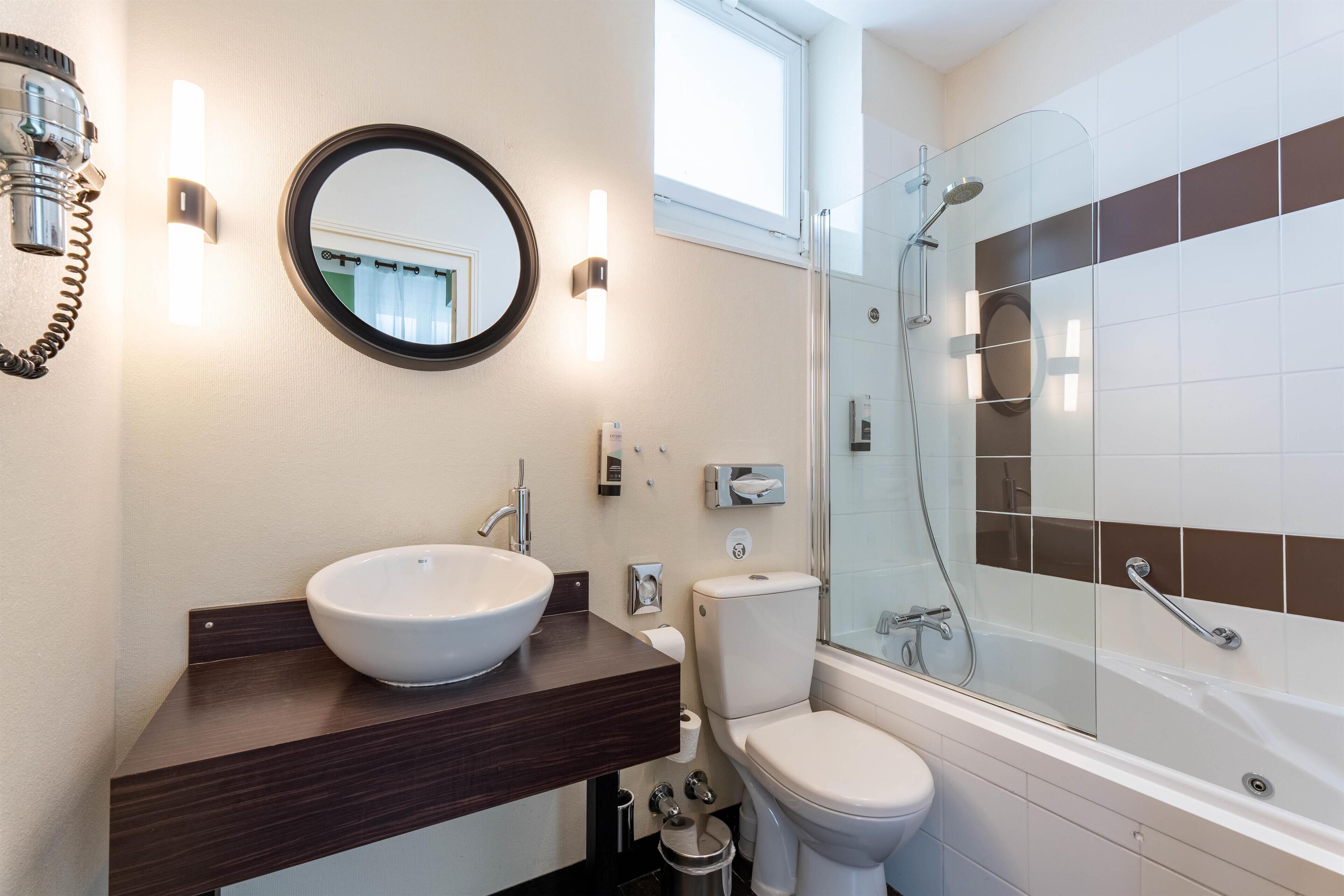 A modern bathroom with a round mirror, a vessel sink, a toilet, and a glass-enclosed shower.