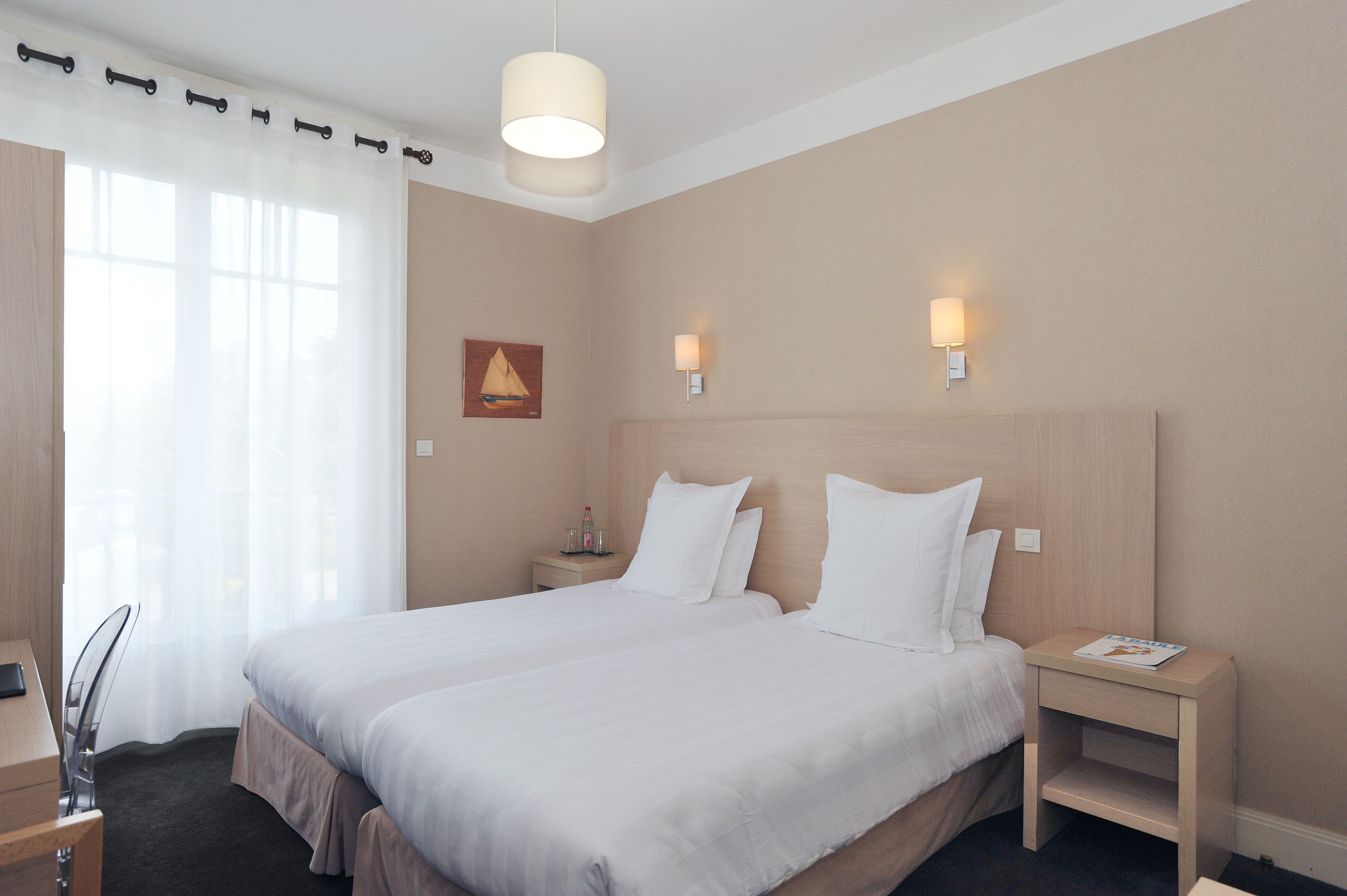 Une chambre d’hôtel avec un grand lit, deux tables de chevet, une petite table, une chaise, un bureau et une fenêtre avec des rideaux légers.