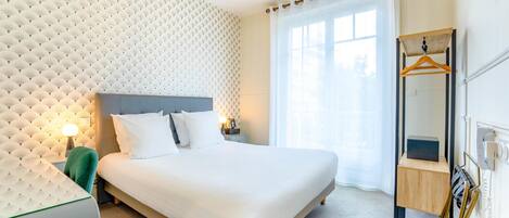 Una habitación de hotel moderna con una cama grande, un cabecero con textura, una mesita de noche con lámpara y un espejo con marco de madera.
