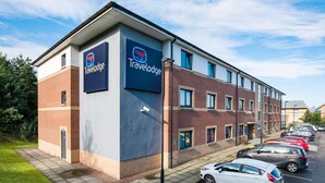 A Travelodge hotel with a brick facade, multiple windows, and a parking lot with several cars. - Travelodge Dunfermline (Dunfermline)