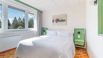 A hotel room with a large bed, a window with curtains, a painting on the wall, and a radiator under the window.