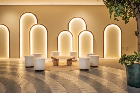 A lobby with a round wooden table surrounded by white cushioned stools, a potted plant, and illuminated wall panels. - IFA Schöneck, Hotel & Ferienpark (Schoeneck)
