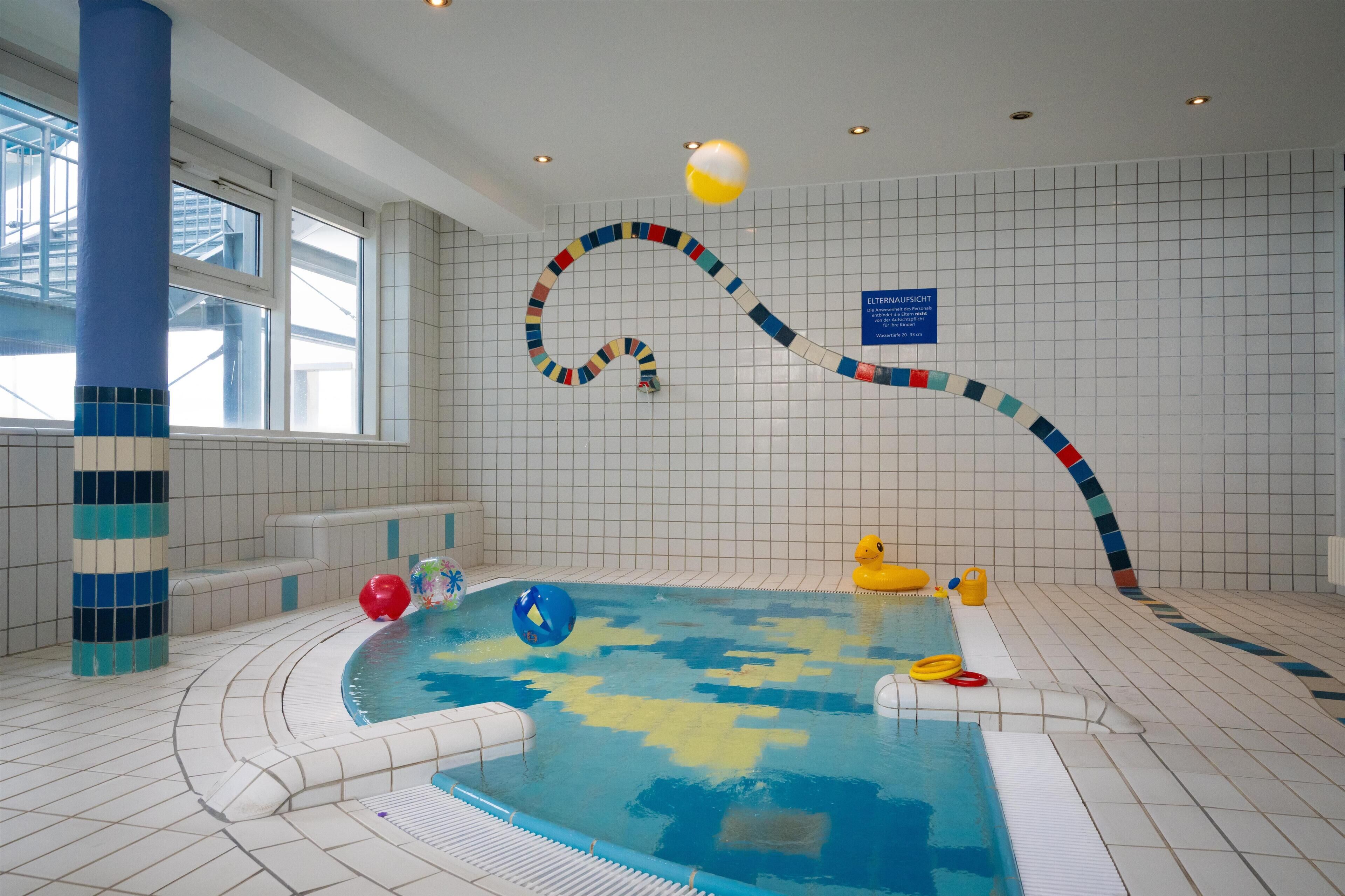 Una piscina interior con un tobogán acuático colorido y serpenteante, suelo con baldosas azules y blancas, y vistas al exterior a través de grandes ventanales.
