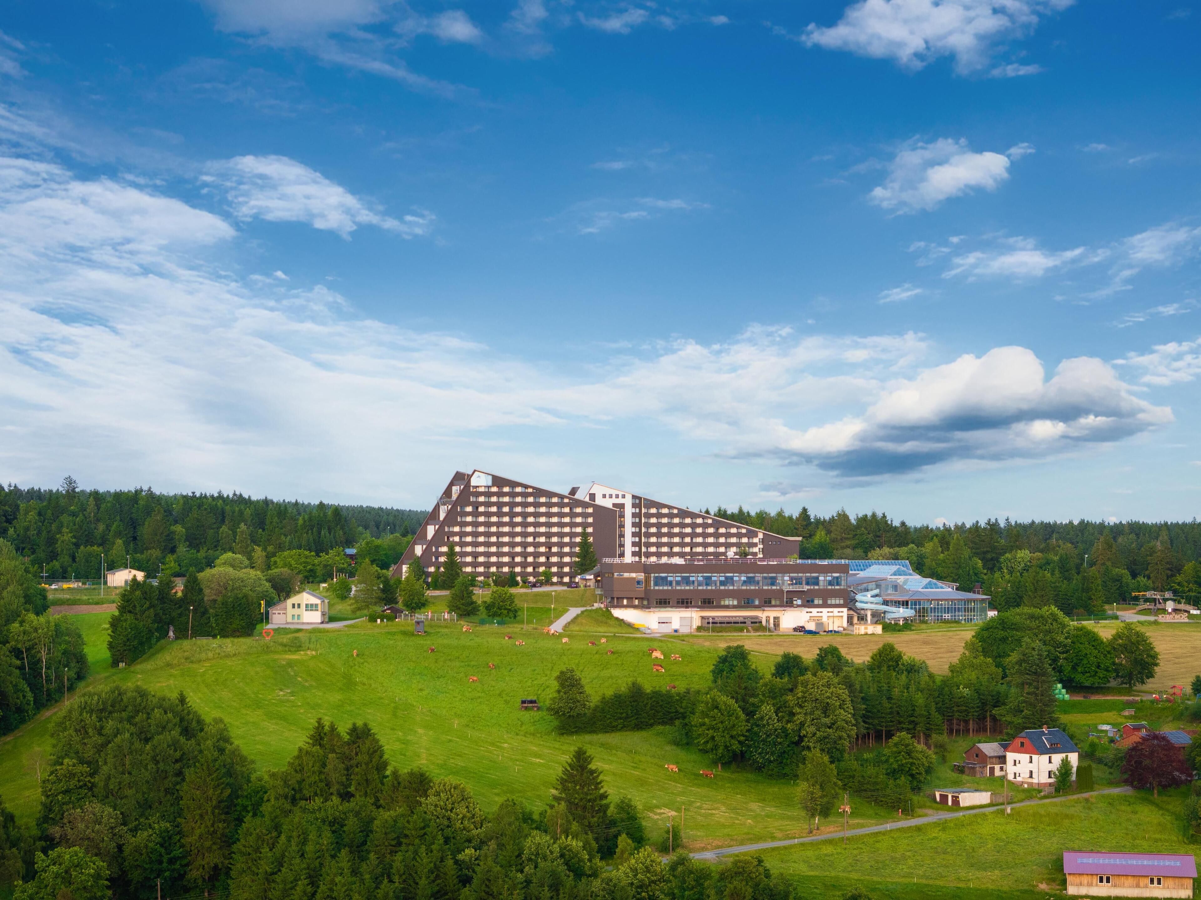 Un hotel grande con varios pisos y techo plano, rodeado de campos verdes y árboles.