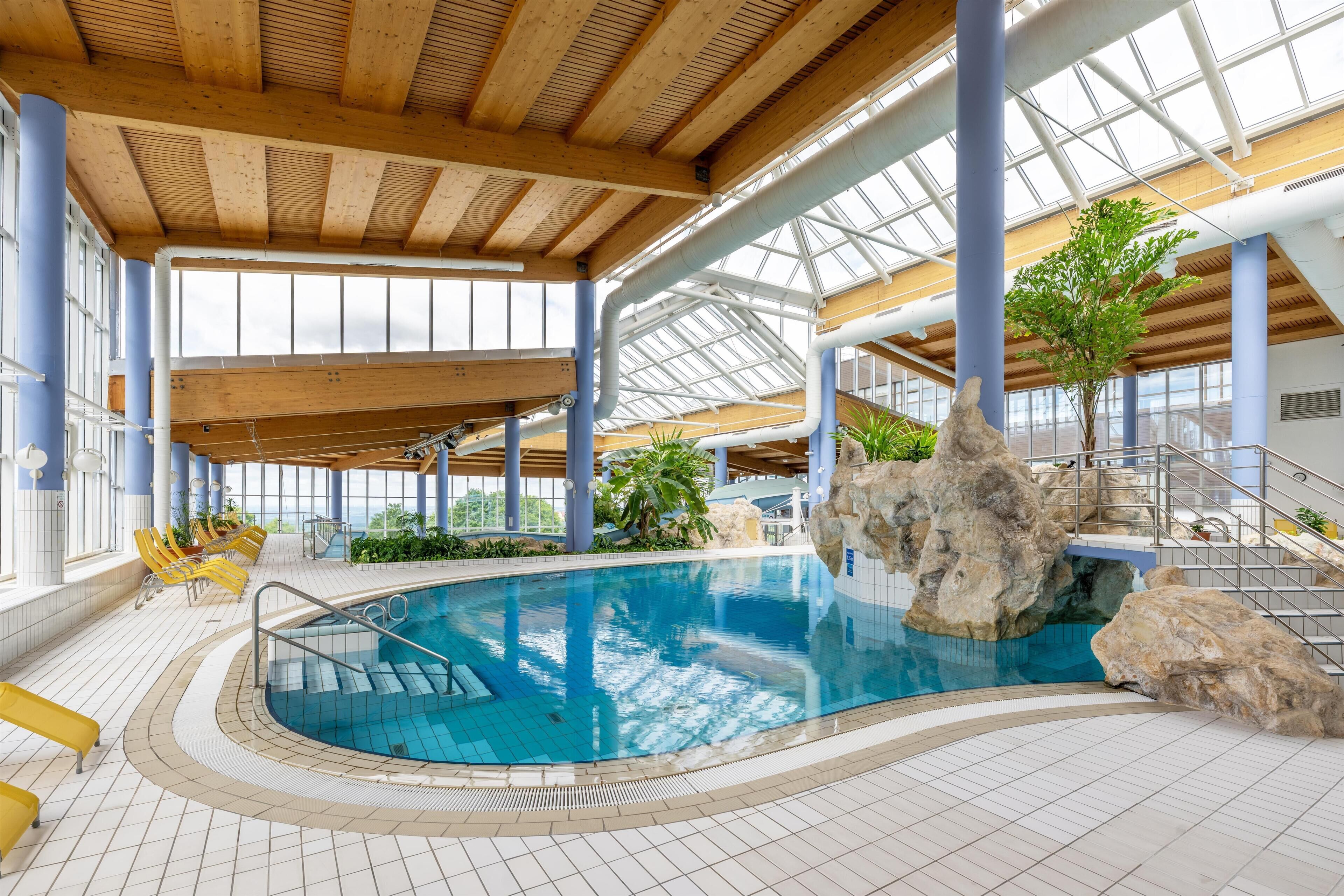 An indoor swimming pool with a wooden ceiling, large windows, and a rock feature.