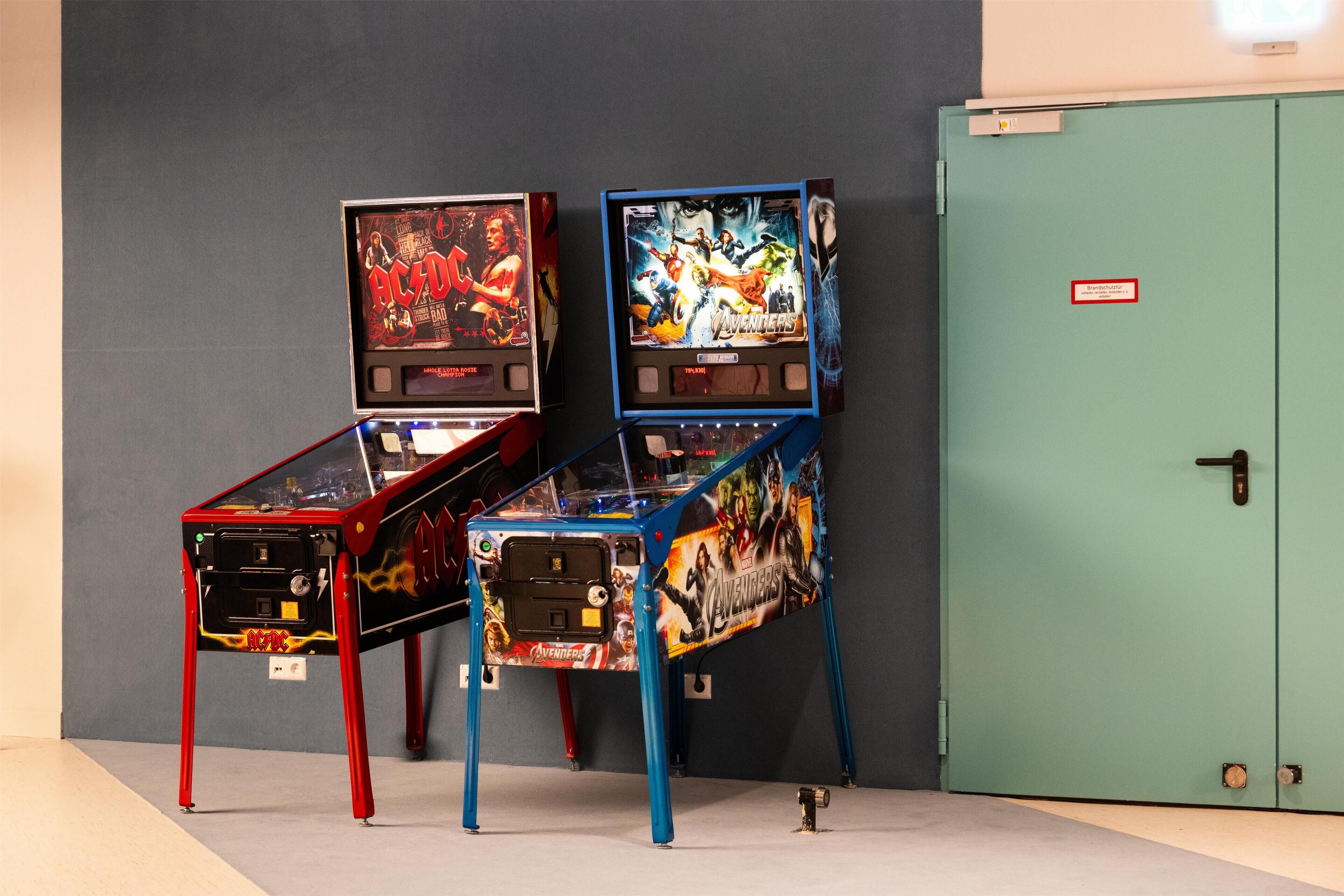 Two arcade machines with 'AC/DC' and 'Avengers' themes, placed against a grey wall with a green door.