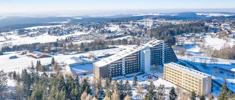 Um hotel alto com vários andares, cercado por paisagens cobertas de neve e florestas densas.