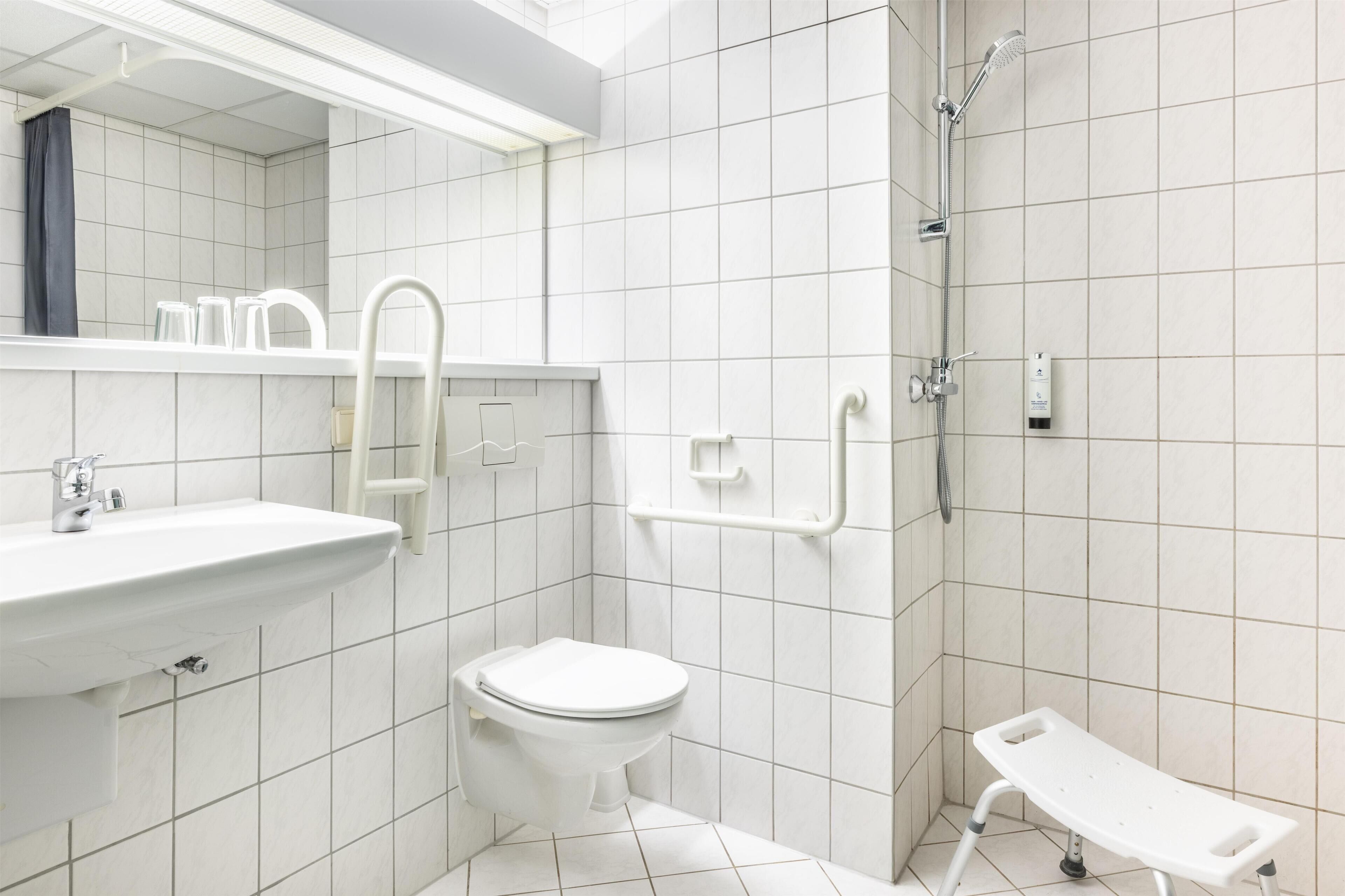 Baño con lavamanos, inodoro y ducha blancos. Cuenta con barras de apoyo y un banco blanco.