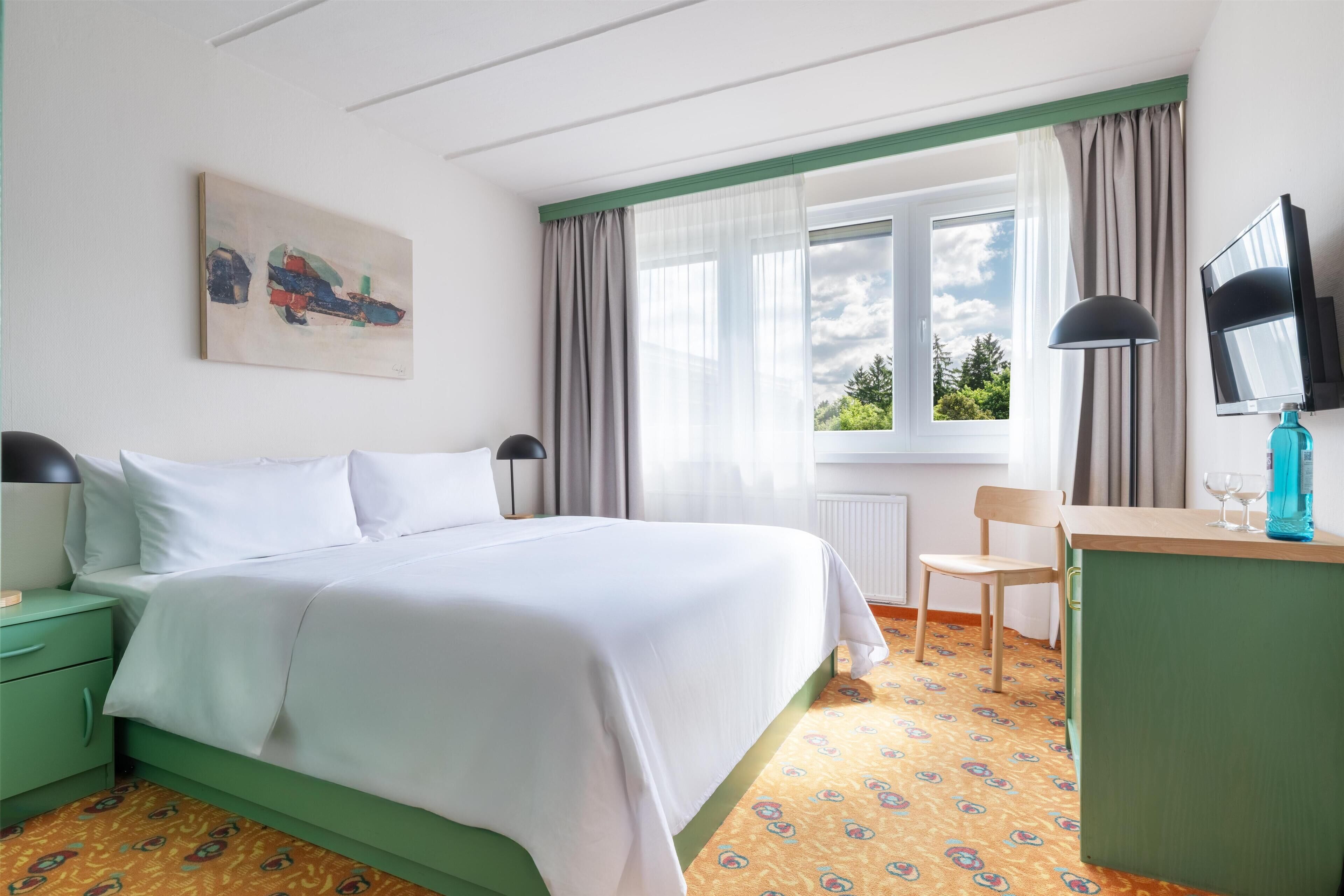 A hotel room with a bed, a desk, a chair, a TV, and a window with curtains.