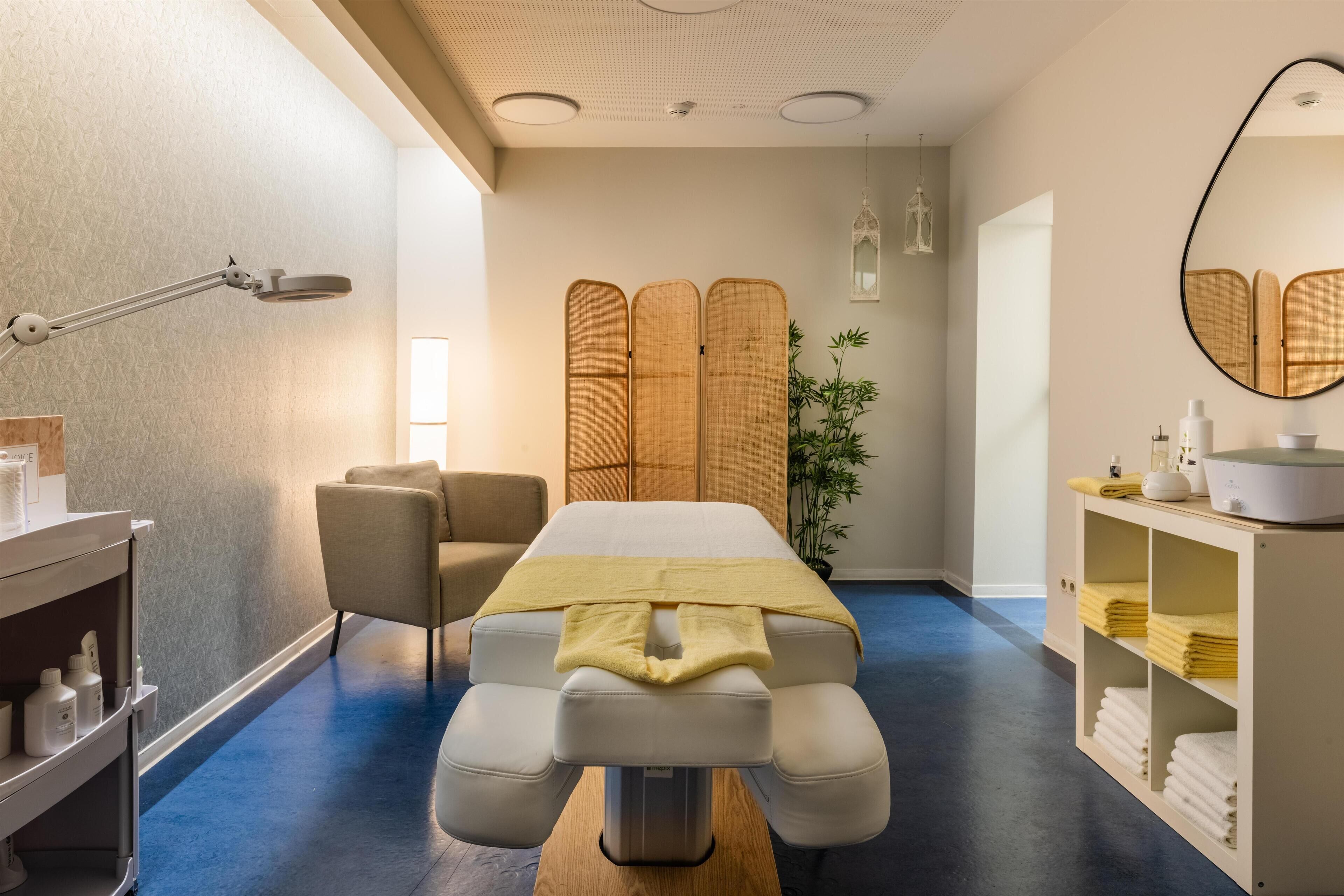 A modern spa room with a massage table, a chair, a shelf with towels, and a mirror on the wall.