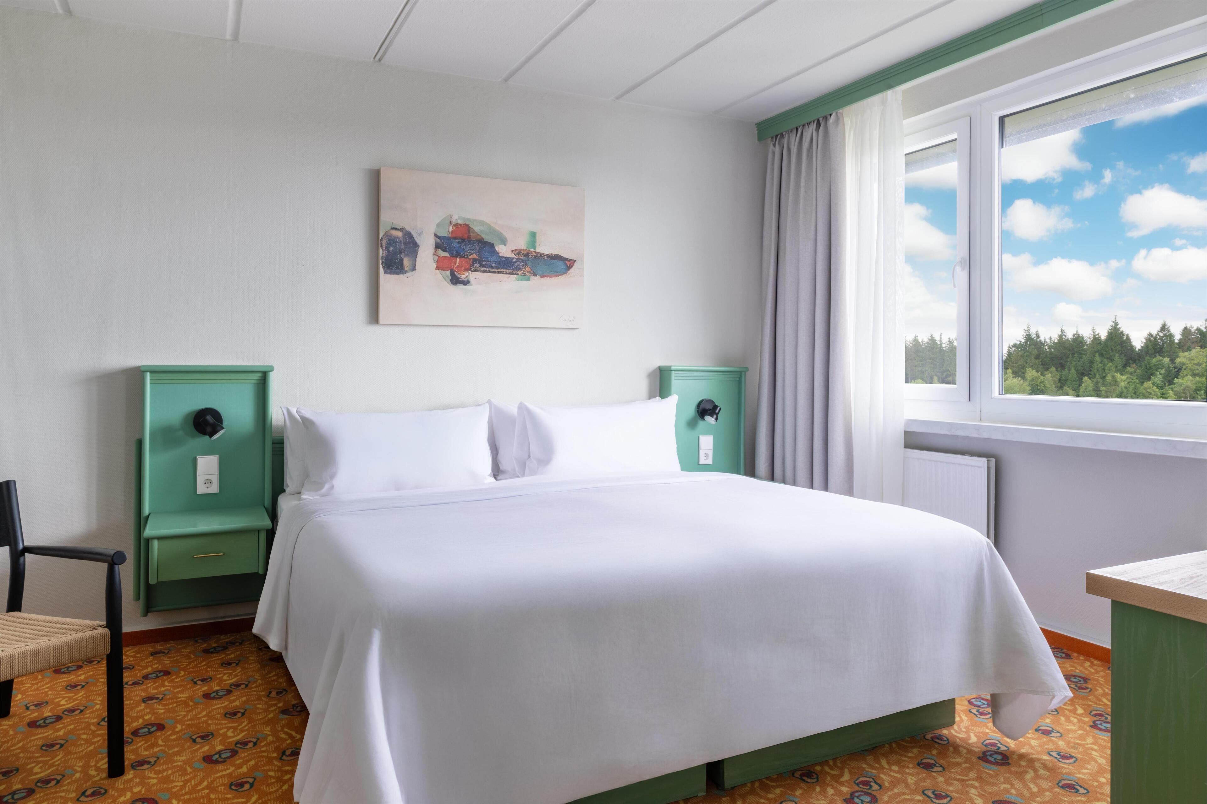 A hotel room with a bed, a chair, a desk, a window with a view of trees, and a painting on the wall.