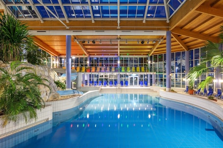An indoor swimming pool with a glass ceiling, blue chairs, and potted plants.