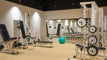 Un gimnasio con diversos equipos de ejercicio, incluyendo máquinas de pesas y pesas libres, dispuesto en una sala amplia con techo alto e iluminación abundante.