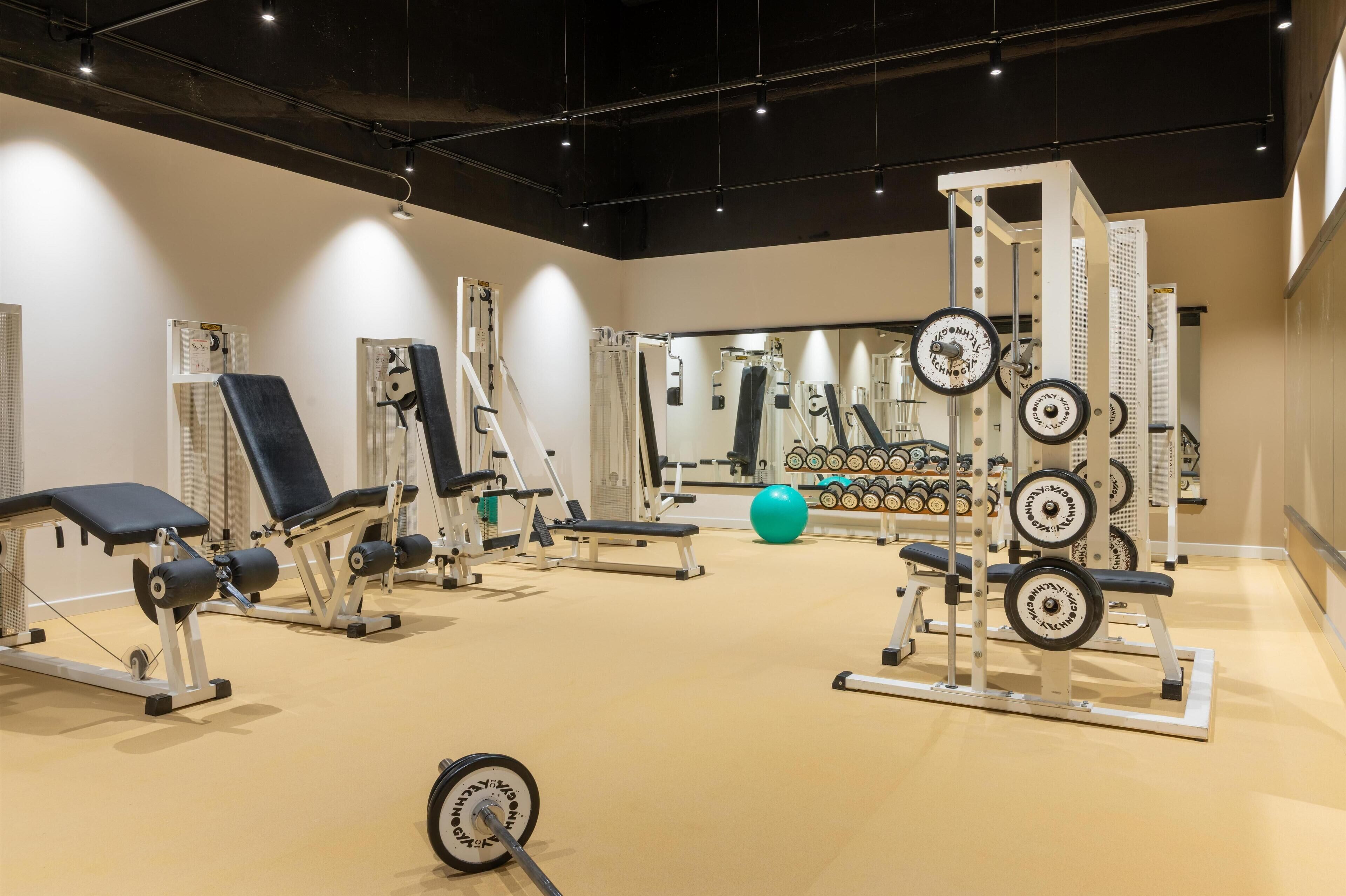 Ein Fitnessstudio mit verschiedenen Trainingsgeräten, darunter Kraftgeräte, Kurzhanteln und einer Spiegelwand.