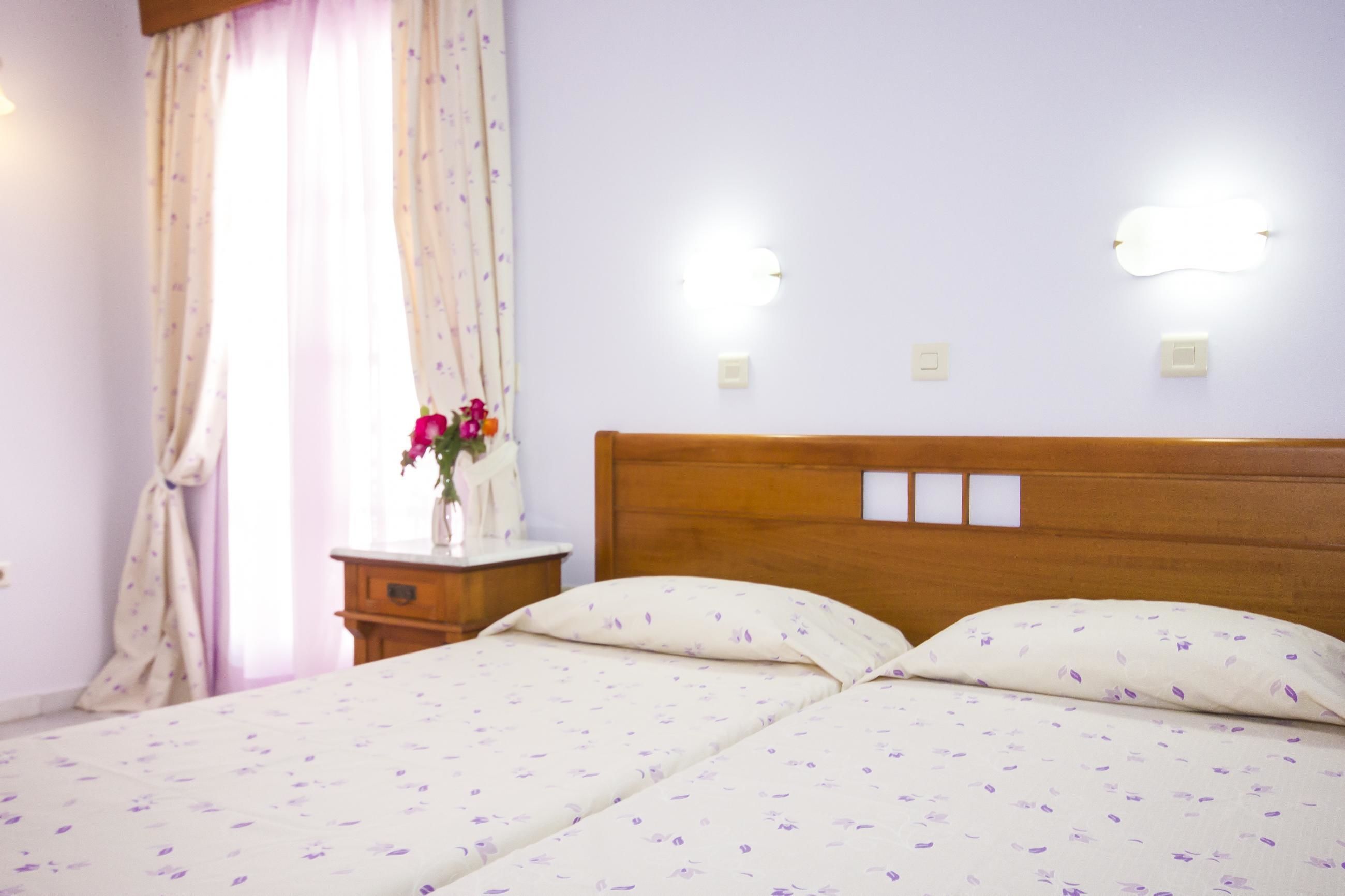A hotel room with a wooden headboard, two beds with floral bedding, a nightstand with a vase of flowers, and wall-mounted lights.