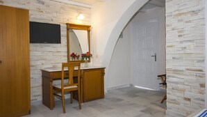 A room with a wooden desk, a chair, a TV mounted on a stone wall, and a door with a stone wall on one side. - Naxos Holidays (Naxos)