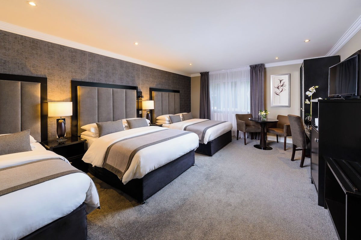 A hotel room with three beds, a dining table with chairs, a TV, and a desk with a lamp.