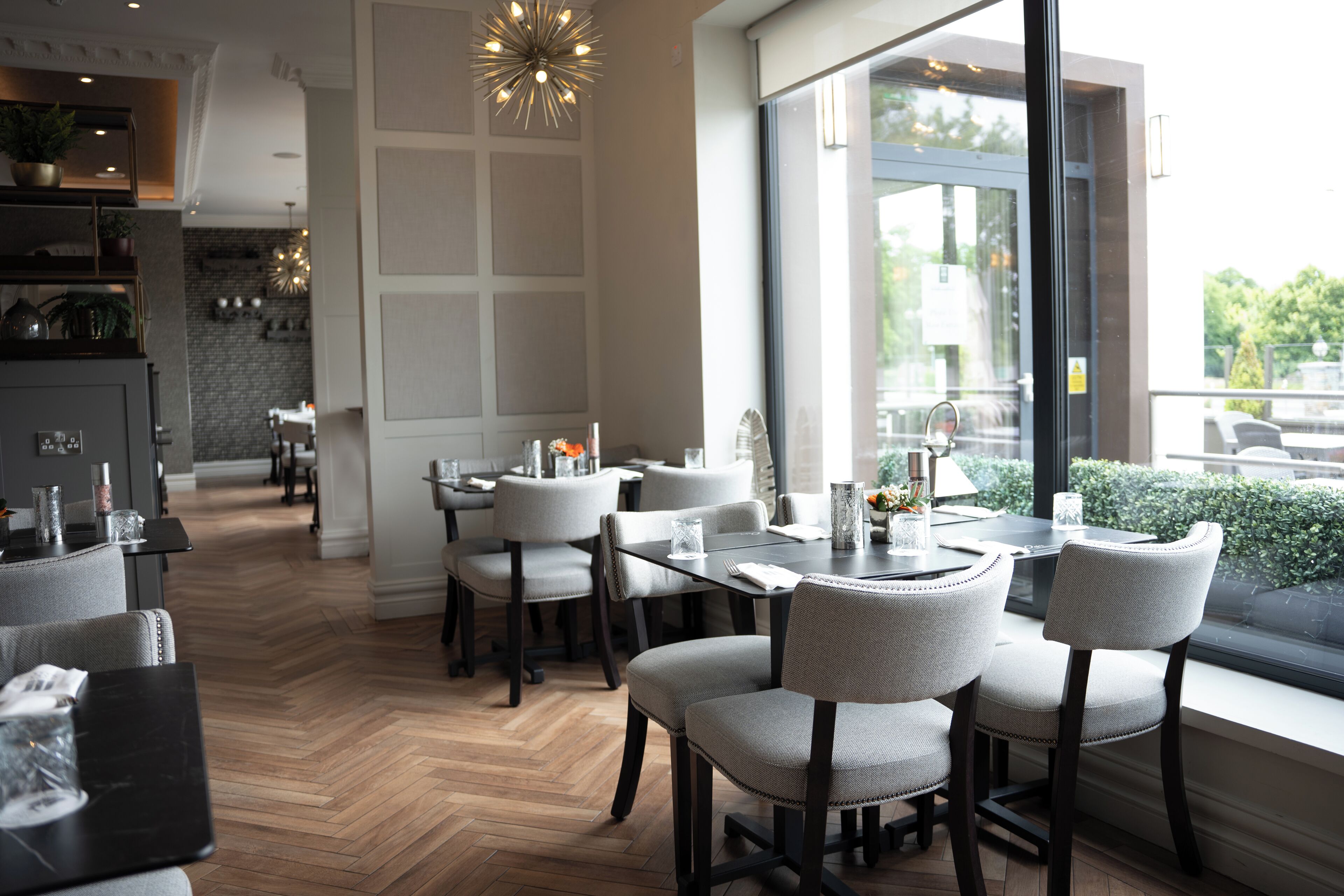 A modern dining area with a herringbone wood floor, grey upholstered chairs, and a large window offering a view of greenery.
