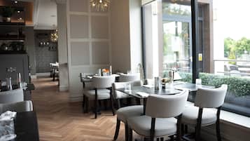 A modern dining area with a herringbone wood floor, grey upholstered chairs, and a large window offering a view of greenery.