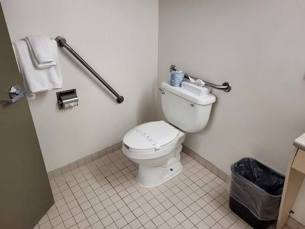 Un baño estándar con inodoro, barra de apoyo, contenedor de basura y dispensador de toallas.